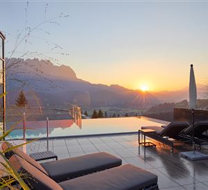 Ein wunderschöner Poolbereich mit Liegen und Blick auf die Berge. Die Sonne geht hinter den Bergen unter und taucht die Szene in warmes Licht.
