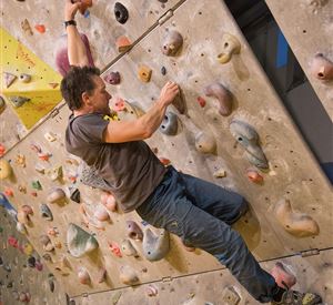 Ein Kletterer, der an einer Kletterwand mit verschiedenen Griffen unterwegs ist. Die Wand ist bunt und hat zahlreiche kleine Haltegriffe.