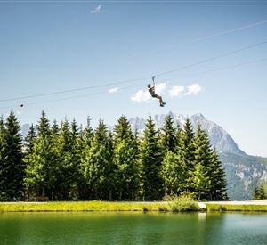 Een avonturier zoeft met de zipline over een helder meer. Op de achtergrond rijst een berglandschap op met dichte bossen.
