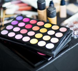 Een make-up palette met veel kleurrijke oogschaduwkleuren ligt op een tafel. Op de achtergrond zijn meer make-up producten zichtbaar.