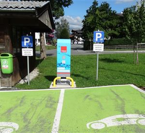 Een parkeerplaats voor elektrische voertuigen met een oplaadstation en zichtbare markeringen. Op de achtergrond zijn er meer parkeerplaatsen en een groen gebied.