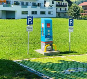Een parkeerplaats met een automaat voor parkeerbewijzen staat op een groene weiland. Op de achtergrond zijn woongebouwen te zien.
