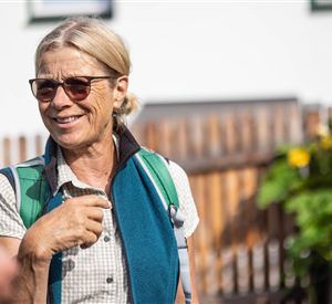 A smiling woman is wearing sunglasses and has a backpack. In the background, green plants and a wooden wall can be seen.