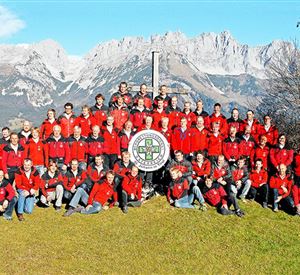 A large group of people in red jackets stands on a meadow in front of majestic mountains. They are posing around a shared sign in the middle of the picture.