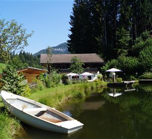 Een rustige vijver met een boot en omringende groene weilanden. Op de achtergrond is een houten huis en enkele parasols te zien.