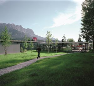 Een modern gebouw in een groene omgeving met bergen op de achtergrond. Een man loopt langs een pad dat door het gras leidt.