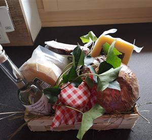 Een mooie geschenkorf met verschillende kaassoorten, een fles azijn en verse kruiden. De korf is versierd met stro en zorgt voor een uitnodigende sfeer.
