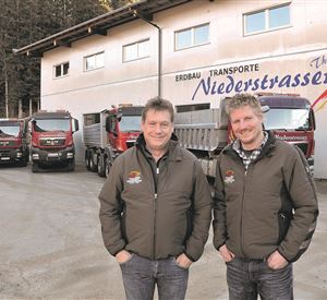 Twee mannen staan voor een vrachtwagenbedrijf met verschillende vrachtwagens op de achtergrond. Het gebouw heeft de inscriptie "Niederstrasser Transporte".