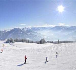 A snow-covered landscape with skiers and a clear, blue sky. In the background, majestic mountains are visible.