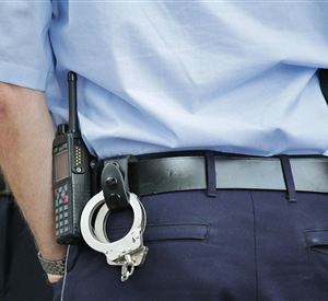 Een politieagent van achteren, die handboeien en een portofoon aan zijn riem heeft. De uniform is blauw en de focus ligt op de inzetmiddelen.