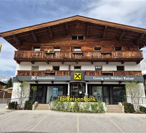 Een moderne bankfiliaal in alpine stijl met houten bekleding. Voor de bank staan gele vlaggen en er is groene beplanting.