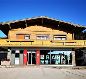 Een modern bankgebouw in alpine stijl met houten bekleding. Op de voorgrond zijn grote vensters met de tekst "BANK" te zien.