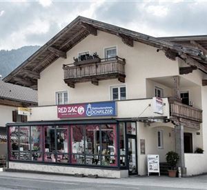 Ein traditionelles Gebäude mit einem Geschäft im Erdgeschoss und Balkonen im Obergeschoss. Die Umgebung zeigt ländliche Architektur und eine bergige Kulisse.