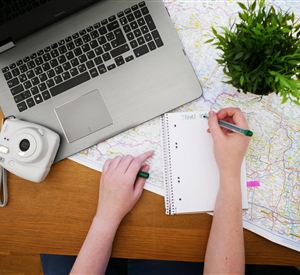 A desk with a laptop, a camera, and a folder. A hand is writing notes on a sheet of paper while a green plant stands in the background.
