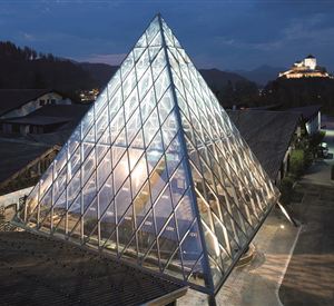 A modern glass pyramid at night, surrounded by traditional buildings. In the background, a castle can be seen on a hill.