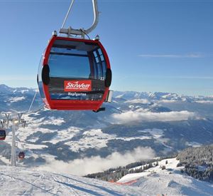 A red gondola hovers over snow-covered mountains. In the background, clouds and additional mountain peaks can be seen.