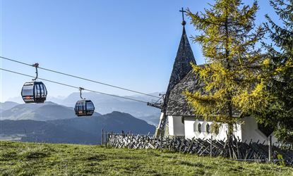 A picturesque mountain landscape with a chapel and cable car cabins. In the background, gentle hills and the Alps are visible.
