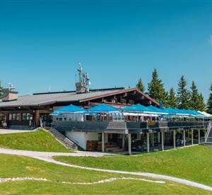 Ein modernes Restaurant in den Bergen mit einer großen Terrasse und blauen Sonnenschirmen. Umgeben von grünen Wiesen und Nadelbäumen unter einem klaren blauen Himmel.