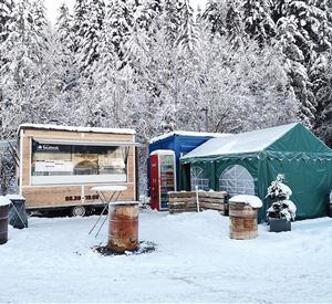 Een verkoopstand en een tent staan in een besneeuwd winterlandschap. Op de achtergrond zijn besneeuwde bomen zichtbaar.