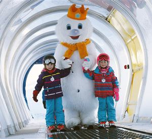 Twee kinderen staan naast een grote sneeuwman in een winterse gang. Ze dragen kleurrijke winterkleding en glimlachen vrolijk.