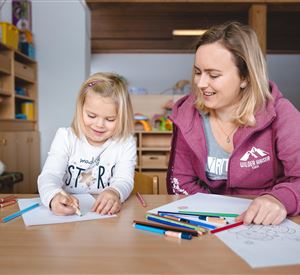 A cheerful girl and a young woman are sitting at a table and drawing. Colorful pens and papers are laid out in front of them.