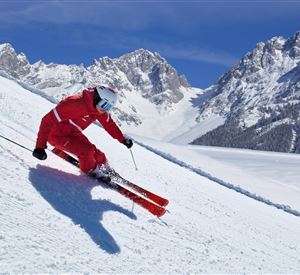 Een skier in het rood gaat een met sneeuw bedekte piste naar beneden. Op de achtergrond zijn majestueuze bergen en een heldere blauwe lucht te zien.