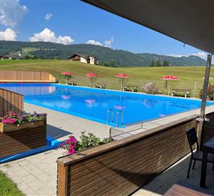 Ein schwimmendes, klares blaues Wasserbecken mit Blumen und Liegestühlen. Im Hintergrund sind grüne Wiesen und Berge sichtbar.