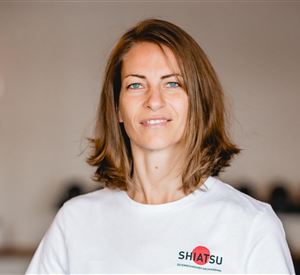 Eine Frau mit braunen Haaren steht lächelnd in einem hellen Raum. Sie trägt ein weißes T-Shirt mit dem Aufdruck "SHIATSU".