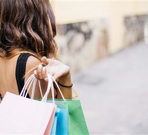 Eine Person trägt mehrere Einkaufstaschen in verschiedenen Farben. Im Hintergrund ist eine schlichte Straße zu sehen.