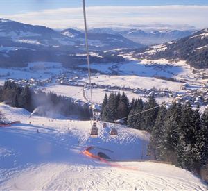 A picturesque snow landscape with a ski lift that rises above green fir trees. In the background, majestic mountains and a small town are visible.