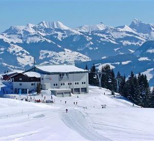 Eine schneebedeckte Berglandschaft mit einer modernen Berghütte. Im Hintergrund sind majestätische Berge und ein klarer blauer Himmel zu sehen.