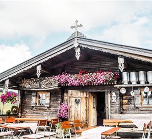 Een traditioneel oud houten huis met kleurrijke bloemen en vriendelijke zitplaatsen buiten. De lucht is gedeeltelijk bewolkt en de sfeer is gezellig.