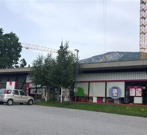 Ein modernes Gebäude mit bunten Fassaden und einem Parkplatz im Vordergrund. Im Hintergrund sind Bäume und ein grüner Hügel zu sehen.