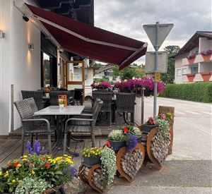 Ein gemütlicher Außenbereich eines Restaurants mit Tischen und Stühlen. Bunte Blumenampeln und dekorative Pflanzen verleihen dem Platz eine einladende Atmosphäre.