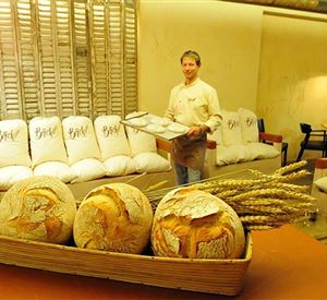 Ein Bäcker präsentiert frisches Brot in einem gemütlichen Café. Im Vordergrund sind dekorativ angeordnete Brote und Deko-Ähren zu sehen.