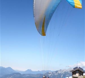 Ein Paraglider startet von einem bewaldeten Hang. Im Hintergrund sind Berge und ein klarer Himmel zu sehen.
