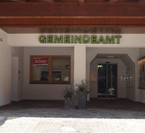 Een gemeentehuis met een uitnodigende ingang. Boven de deur staat "GEMEENTEAMT" in groene letters.