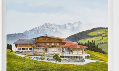 A large, picturesque wooden house in a green hill and mountain landscape. The scene shows clear skies and surrounding forests.