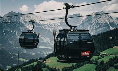 Eine Seilbahn fährt durch eine malerische Berglandschaft. Im Hintergrund sind hohe Berge und grüne Wiesen zu sehen.
