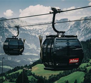 A cable car glides over green meadows and majestic mountains. In the background, tall, snow-capped peaks are visible.