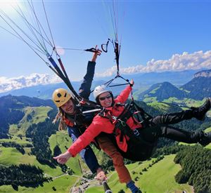 Two people paragliding over a green, hilly landscape. The sky is blue with some clouds.
