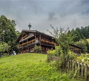 Een traditioneel houten huis omringd door groene weilanden en bomen. Op de achtergrond zijn zachte heuvels en een bewolkte lucht te zien.
