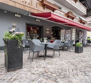 Een gezellige buitenruimte van een café met tafels en stoelen. Rode parasols bieden schaduw, en decoratieve planten zijn ook aanwezig.
