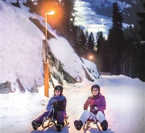 Two people are sitting on a sled in the snowy landscape. It is evening, and the surroundings are illuminated by lights.