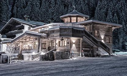A cozy wooden house in the snow, surrounded by tall fir trees. The surroundings are wintry and peaceful.
