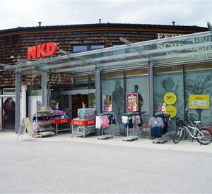 A store of NKD with a modern glass façade. In front of the shop are clothes and advertising boards with offers.