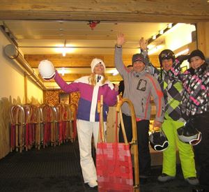 A group of four people in winter clothing stands in a ski room. They are laughing and holding their helmets as well as sleds in their hands.