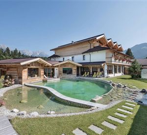 Ein modernes Haus mit Holzarchitektur und großen Fenstern, umgeben von einem gepflegten Garten. Im Vordergrund befindet sich ein natürlicher Pool mit einer geschwungenen Form.