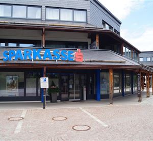 Een modern bankgebouw met de tekst "Sparkasse" in blauwe letters. Het buitengebied is geplaveid en er zijn enkele parkeerplaatsen.