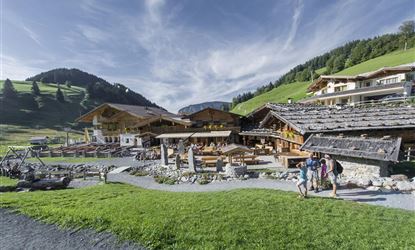 A picturesque alpine inn surrounded by green meadows and gentle hills. Three hikers are walking on a path that leads to the entrance.